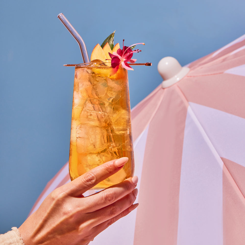 A hand holding White Peach iced tea with peach and flower garnish in a glass with straw. Background is the sky and beach umbrella.