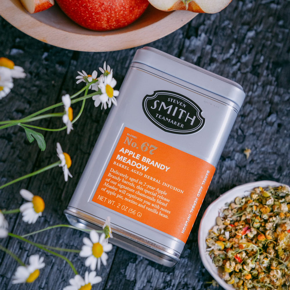 Grey tin of Apple Brandy Meadow with an orange label next to a bowl of loose Meadow tea, a bowl of apples and fresh chamomile flowers.