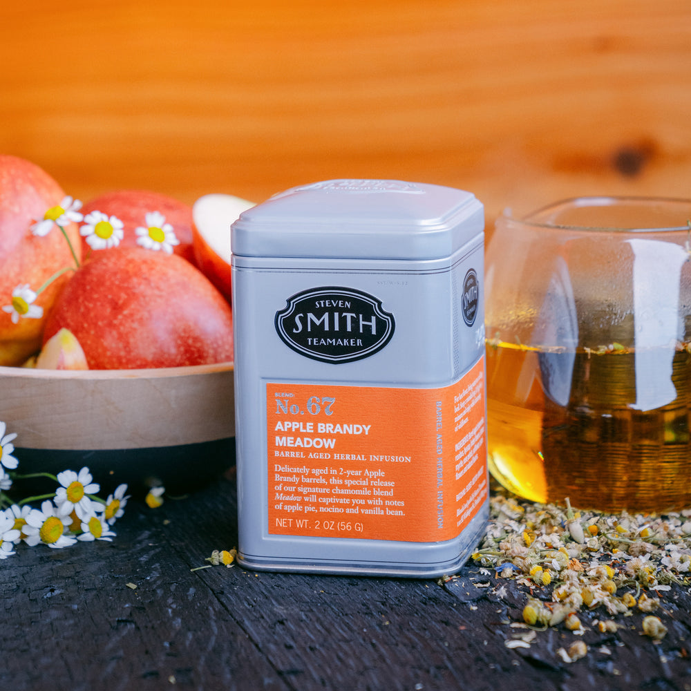 Grey tin of Apple Brandy Meadow with an orange label next to a cup of brewed tea, a bowl of apples and fresh chamomile flowers.