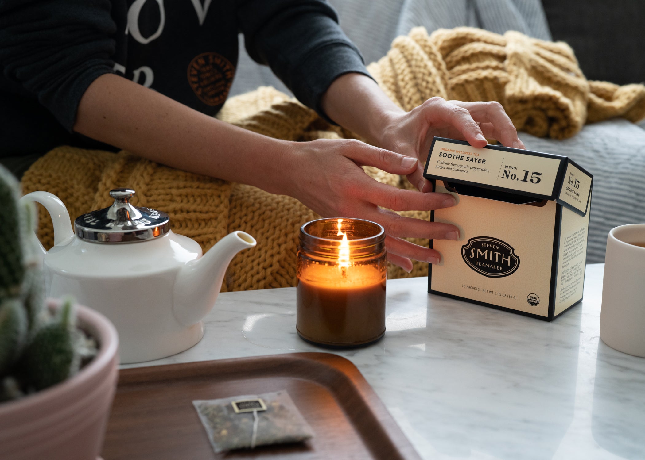 Person on couch with blanket reaching for a box of Soothe Sayer on table with lit candle and teapot.