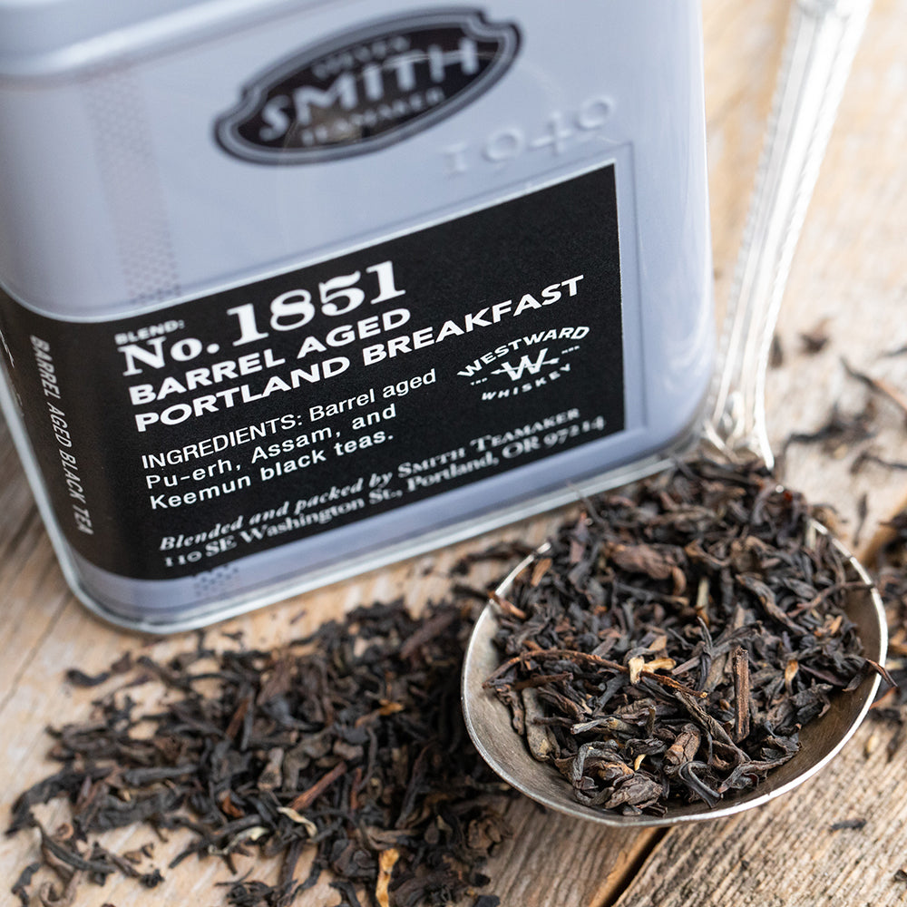 Silver spoon on a wooden table filled with loose black tea next to silver tin with a black label.