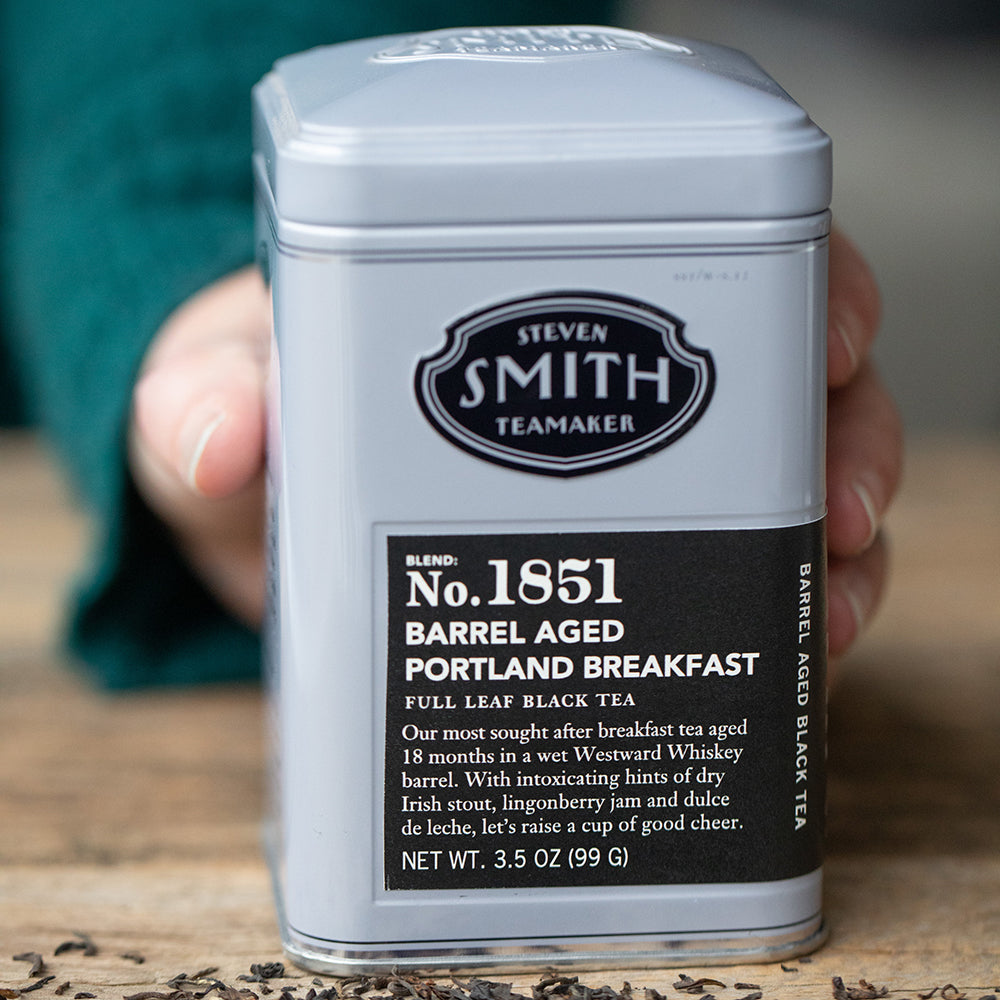 Person holding a silver tin of barrel aged portland breakfast tea with black label on wooden table.