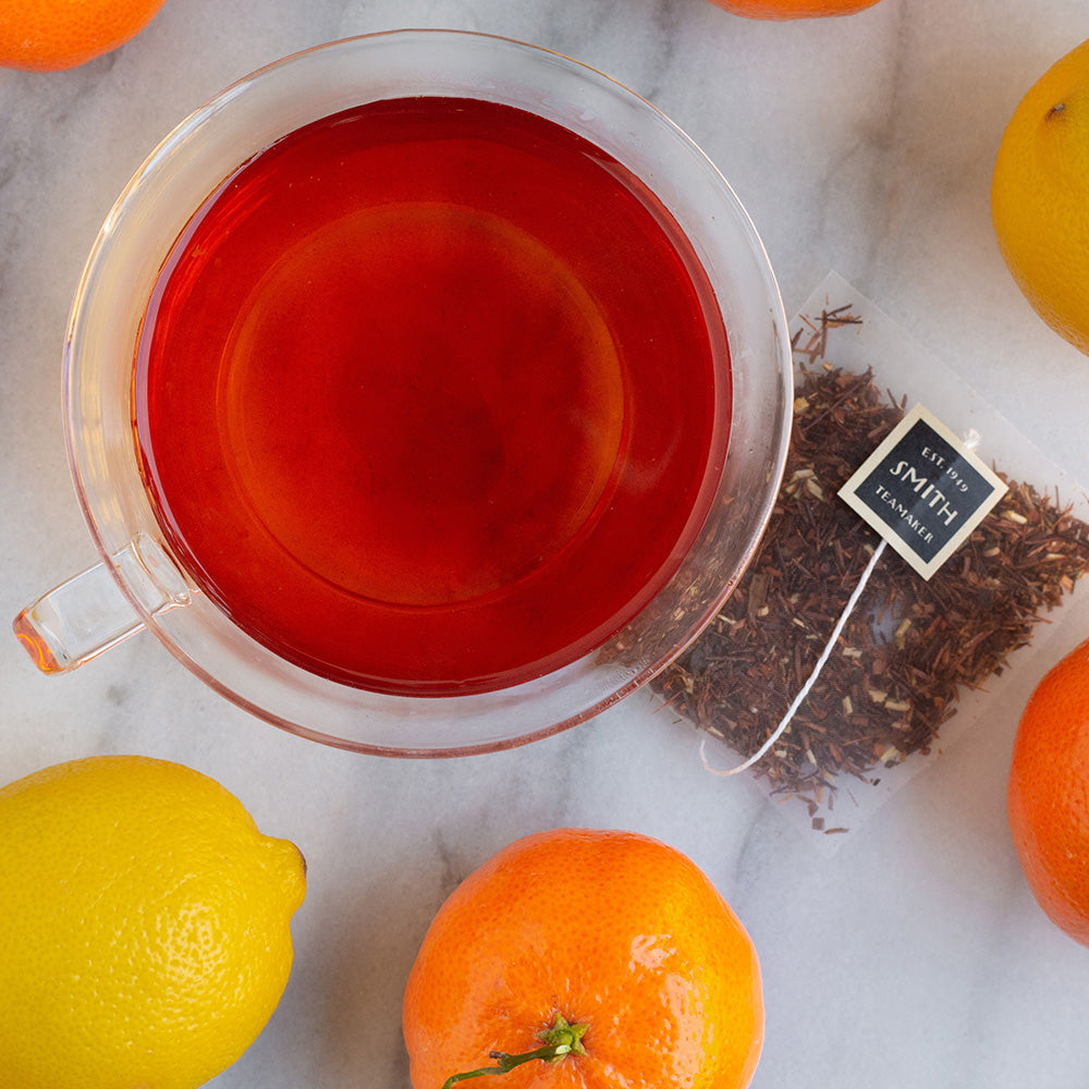Glass cup filled with red nectar herbal tea alongside lemons and oranges.