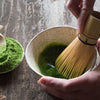 Bamboo matcha whisk with black thread in bowl with green liquid..
